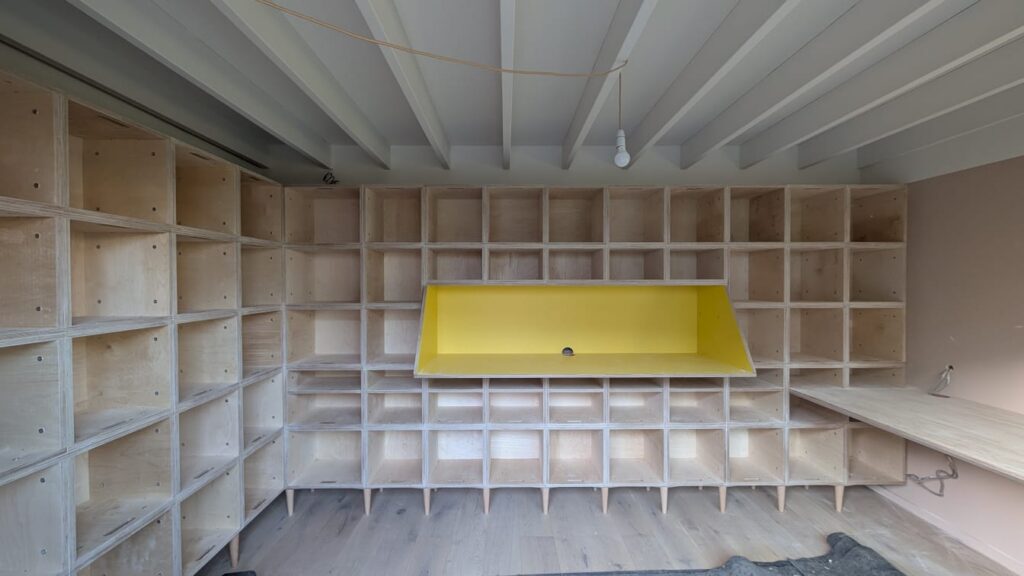 Room with wooden cubby shelves along the walls, a yellow rectangular structure in the centre, exposed ceiling beams, and a hanging light bulb—an industrial-inspired Record Room in East London with a partially finished wooden floor.