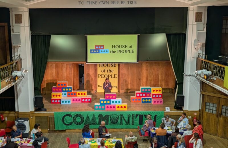 A person speaks on a stage decorated with colourful block props and a “HOUSE OF THE PEOPLE” sign, evoking the spirit of Parallel Parliament; audience members sit at tables below, with “COMMUNITY” displayed across the front of the stage.