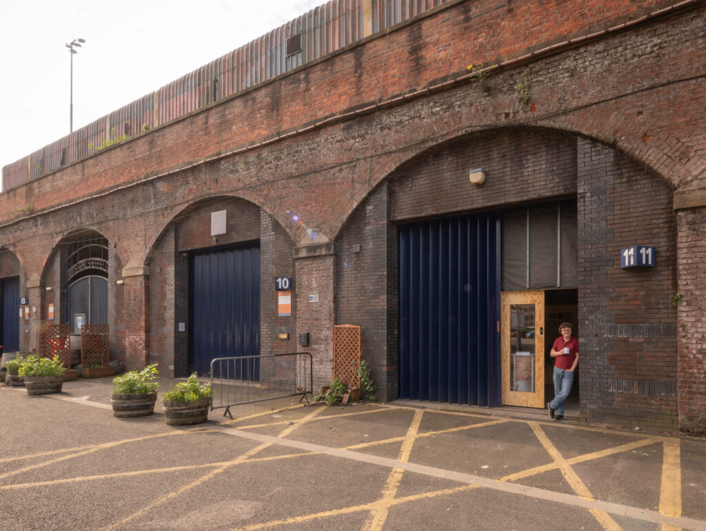 A person leans against the doorway of Dekiln Workshop unit 11, set in a row of brick archways with blue metal doors. Potted plants line the pavement, and parking bays are marked on the ground.