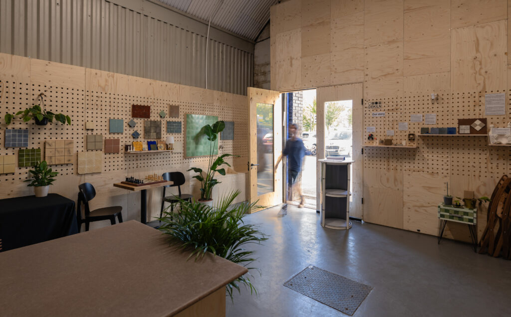 A modern, industrial-style Dekiln Workshop features plywood walls displaying colour samples and plants. Sunlight streams in as a person enters through a glass door. Tables, chairs, and shelves with decor complete the inviting scene.