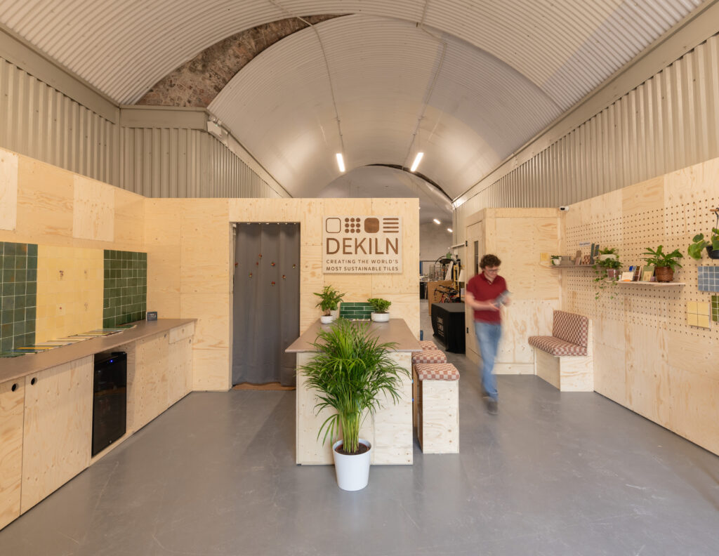 A modern, minimalist Dekiln Workshop features wooden walls and furniture, green tiles on the left, potted plants, and a person walking inside. The sign DEKILN is displayed on the back wall above a central table.