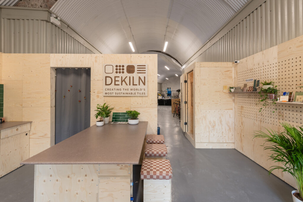 A modern, minimalist Dekiln Showroom with light wood walls, plants, stools around a central table, and a sign reading “DEKILN Creating the world’s most sustainable tiles.” Shelves and a green tile display complete the space.