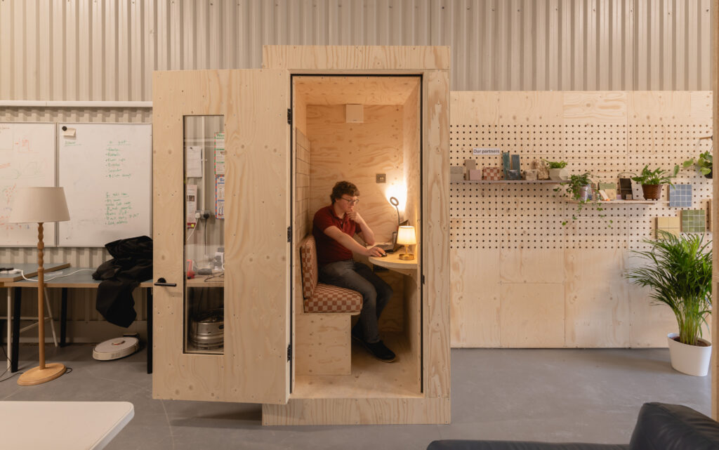 A person sits inside a small wooden Dekiln booth, working at a desk with a lamp. The booth is in a modern office space with light wood walls, plants, shelves, and a whiteboard nearby.