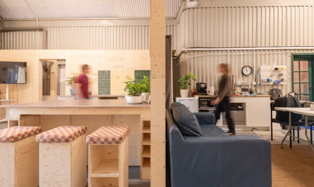 A modern, industrial-style Dekiln Showroom kitchen and lounge space with wooden furniture, a blue sofa, plants, and two blurred figures walking. The walls have a corrugated metal finish, adding character to the open kitchen area.