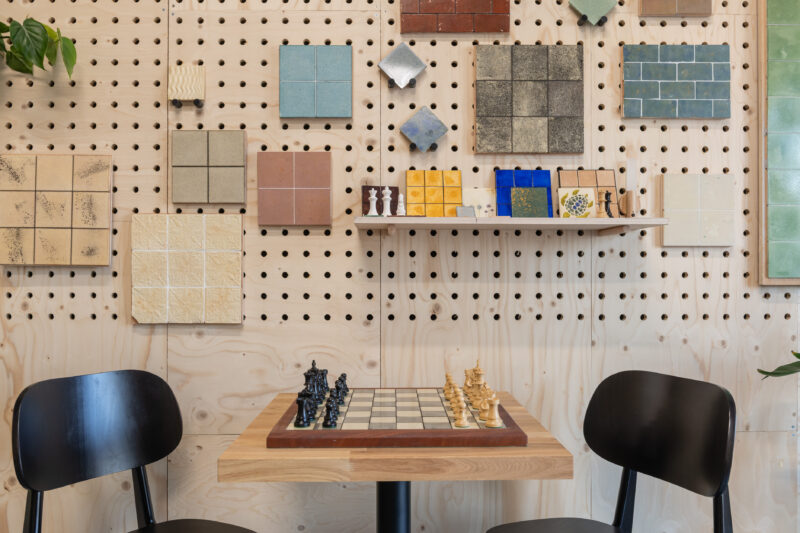 Two black chairs face a square table with a chessboard and pieces. The pegboard wall displays colourful tile samples from the Dekiln Showroom, with some plants partially visible on the sides.