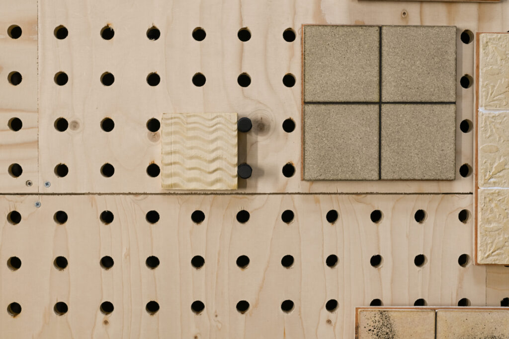 A wooden pegboard with evenly spaced round holes displays various square tiles, including textured beige and patterned tiles, featured in the Dekiln Showroom.