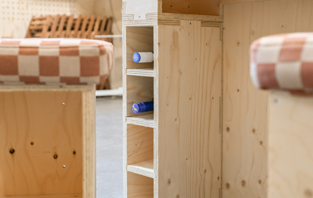 A close-up of wooden furniture from the Dekiln Showroom features a built-in vertical shelf holding three horizontally placed bottles. Cushioned seats with red and white checked fabric are partially visible in the background.