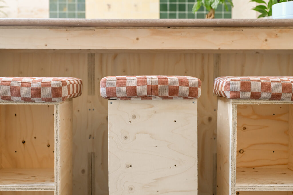 Three wooden bar stools with red and white chequered cushions are lined up in front of a light wooden counter in the bright, modern Dekiln Showroom.