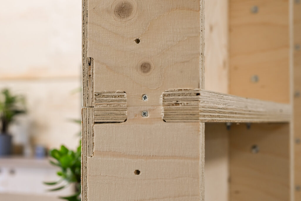 Close-up of interlocking plywood joints secured with screws in a light-coloured wooden structure at Dekiln Workshop, with blurred green plants and shelves in the background.