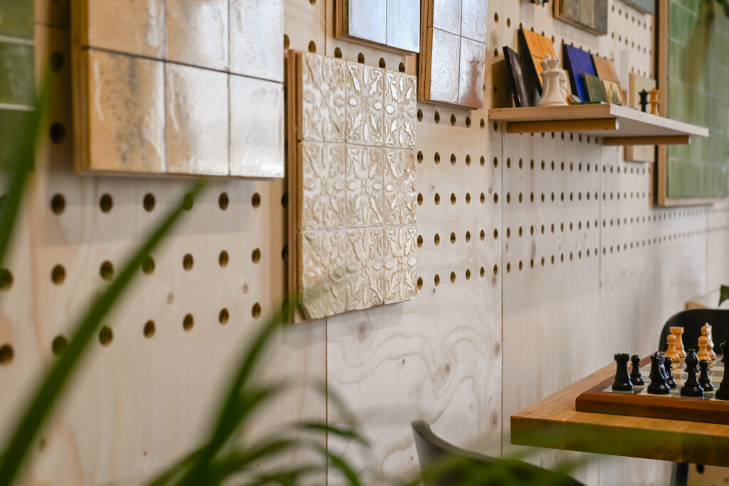 A decorative wall with pegboard panels, shelves holding books and items, and a chessboard on a nearby wooden table create a cosy Dekiln Workshop vibe. Some green plants are partially visible in the foreground.