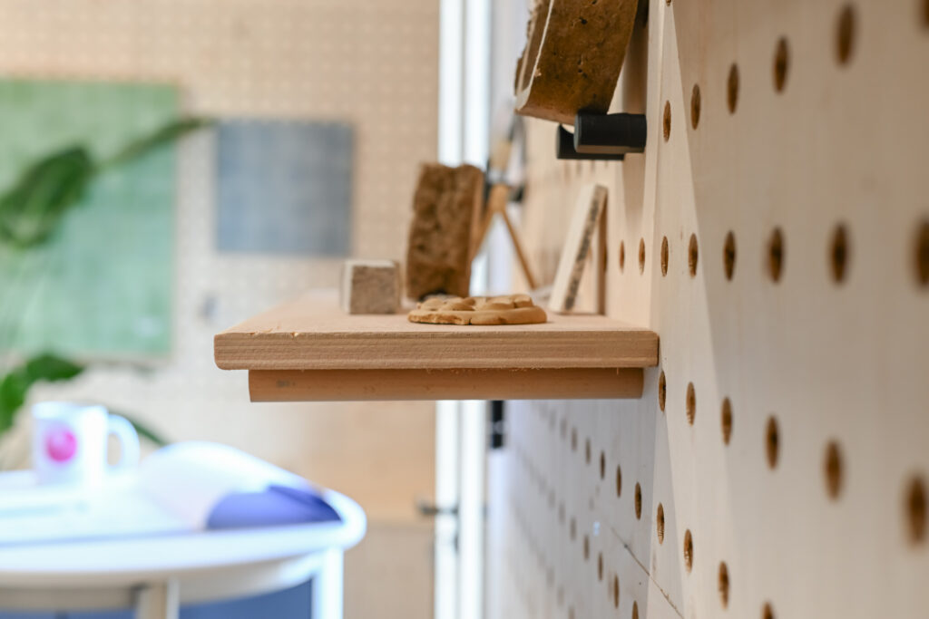 A close-up, side view of a wooden shelf mounted on a pegboard wall at the Dekiln Showroom, holding small objects. The background is blurred, with a chair, mug, and wall art softly visible.