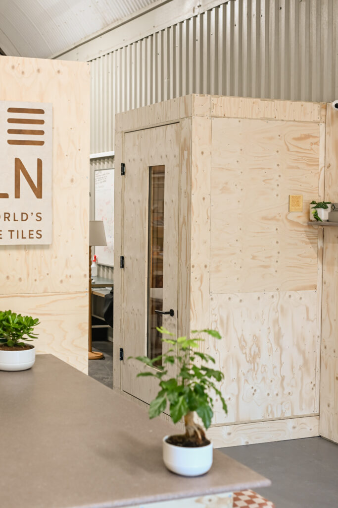 A modern workspace at the Dekiln Showroom features light plywood walls, a small enclosed booth with a glass door, potted plants on the counter, and part of a company sign reading N WORLD’S TILES.