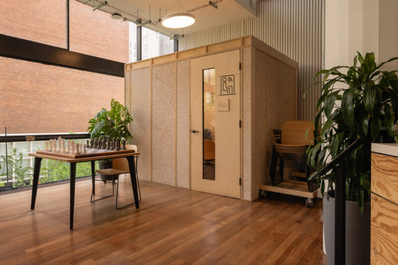 A modern office space in Manchester with wooden floors features a table set up for chess, large green plants from Sustainable Ventures, and a small enclosed meeting room with glass panels and a door. Natural light pours in through tall windows.