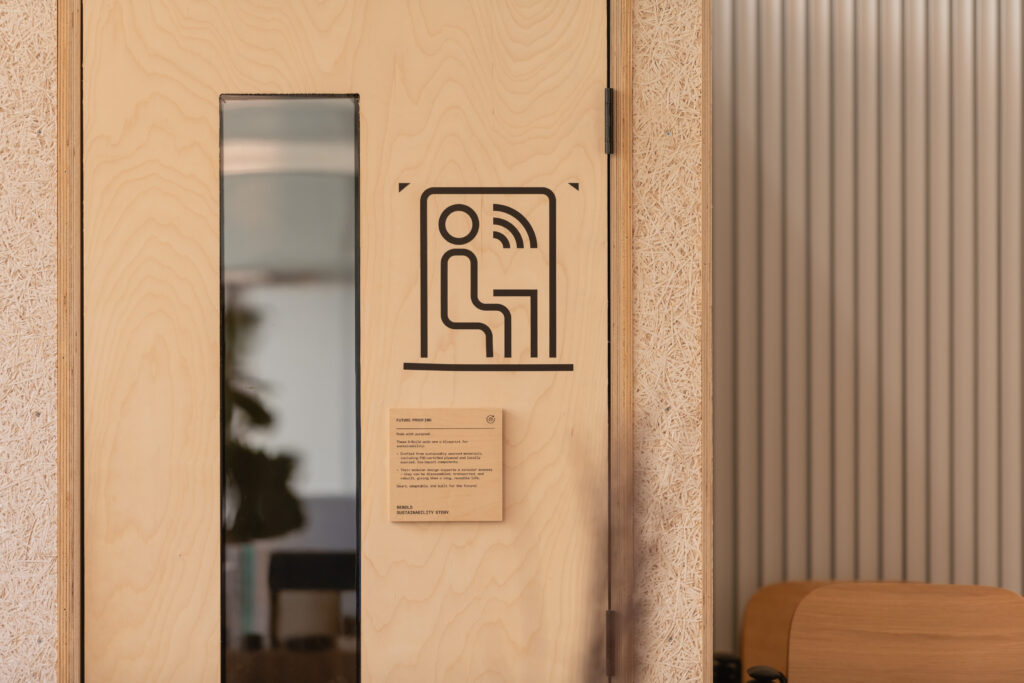 A light wooden door with a narrow vertical window and a black icon showing a person sitting and sound waves, likely at Sustainable Ventures in Manchester. Below the icon is a small plaque. The right side shows part of a corrugated wall and a chair.