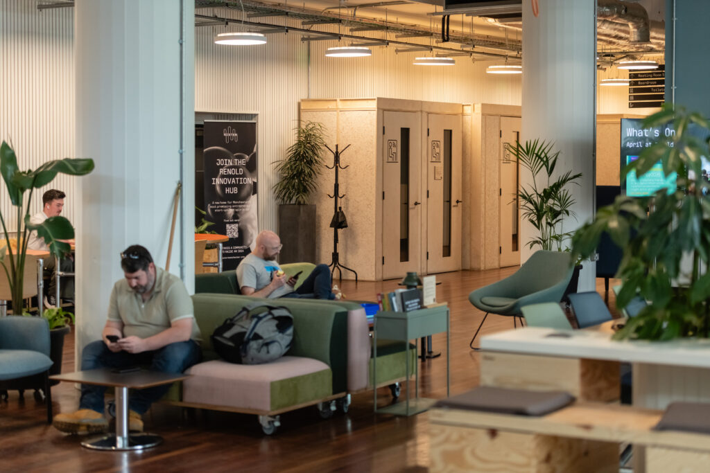 A modern coworking space by Sustainable Ventures in Manchester with people on sofas and chairs using laptops and mobile phones. The area features plants, wooden floors, and doors in the background, creating a relaxed and productive atmosphere.