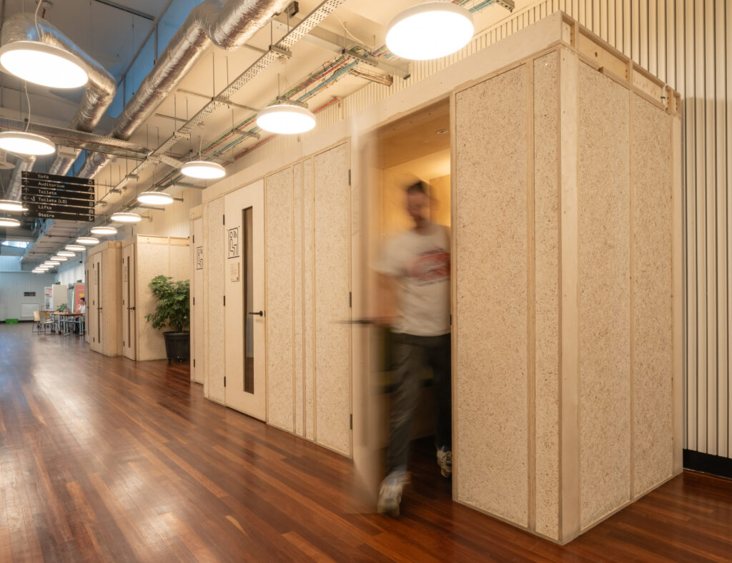 A person walks out of a modern, private booth in Sustainable Ventures, a sustainable business hub in Manchester. The brightly lit corridor features wooden floors, white walls, exposed pipes on the ceiling, and motion blur as the door opens.