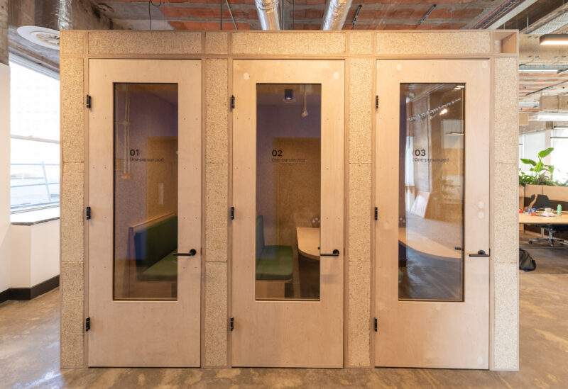 Three small timber U-Build meeting pods with glass doors and numbers 01, 02, and 03 stand side by side in Sustainable Ventures’ modern Manchester office, featuring exposed ceilings, concrete floors, and visible desks to the right.
