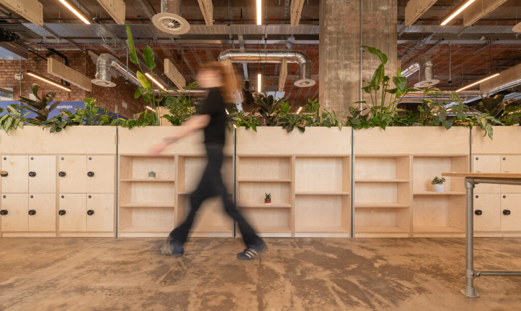 A person in motion walks past wooden shelves and lockers in the Manchester workspace of Sustainable Ventures, featuring exposed pipes, plants, and a concrete pillar—a hub for modern industrial design and sustainable business innovation.