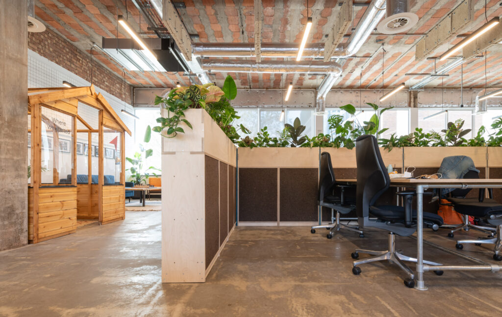 Modern open-plan office in Manchester with an industrial-style ceiling, indoor plants on dividers, ergonomic chairs around a table, and a small wooden structure in the corner—ideal for a sustainable business like Sustainable Ventures. Large windows provide natural light.