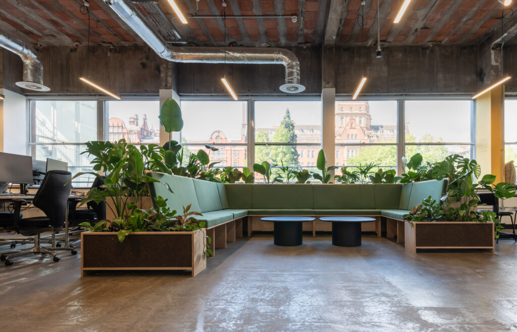 Modern office lounge area with green sectional seating by Sustainable Ventures, surrounded by potted plants, large windows filling the Manchester space with natural light, exposed ceiling pipes, and round black coffee tables in the centre.