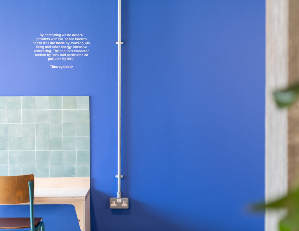 A turquoise-tiled table with a wooden chair sits against a vibrant blue wall in Manchester. A metal conduit runs vertically, and white text about Sustainable Ventures and sustainable business tile production is displayed above the table.