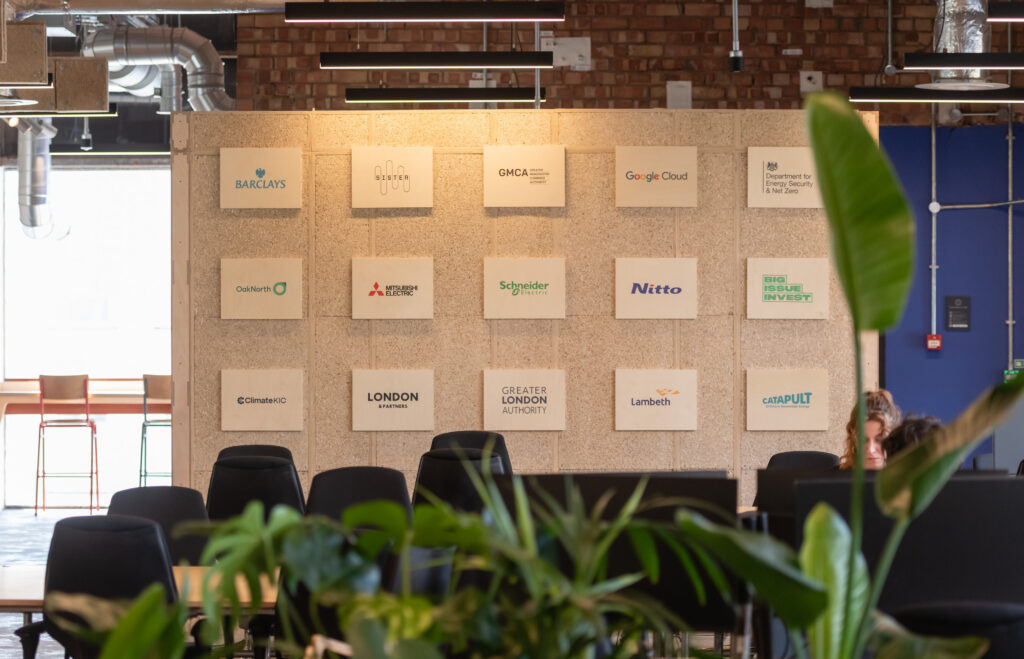 A modern Manchester office space with a wall displaying various company logos, including Barclays, Google Cloud, and Sustainable Ventures. Green plants and empty chairs are visible in the foreground.