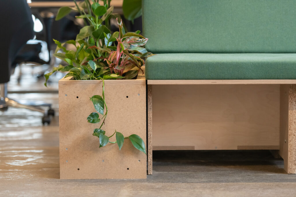 A close-up of a modern bench with a green cushion and an attached wooden planter box filled with leafy plants, set on a concrete floor in a Manchester office space by Sustainable Ventures.