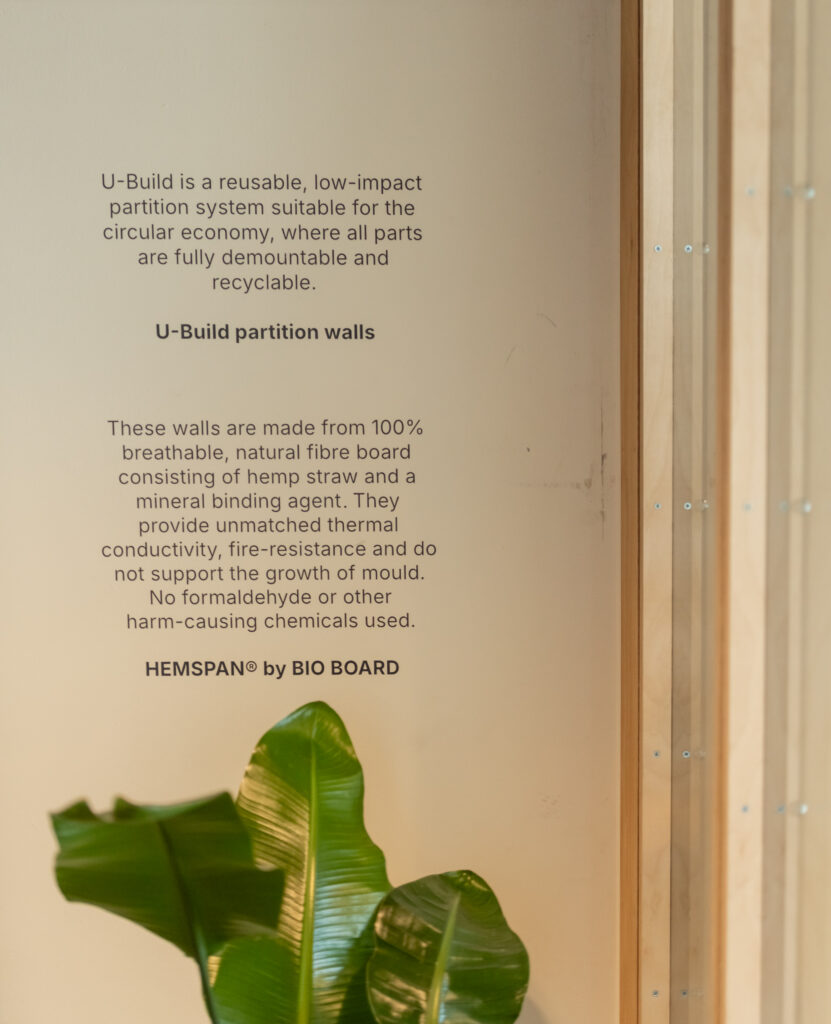 Text on a light-coloured wall at Sustainable Ventures, Manchester, details U-Build’s reusable partition walls—crafted from 100% breathable, natural fibre board with hemp straw and a mineral binder. Nearby, a leaf’s tip and wooden beams are visible beside the text.