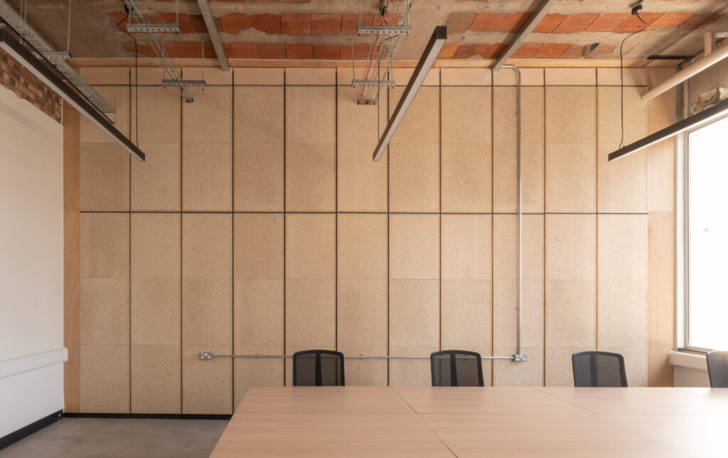 A modern office meeting room in Manchester featuring a long wooden table, five black mesh chairs, an exposed ceiling with pipes and bricks, and a large window letting in natural light—perfect for sustainable business discussions.