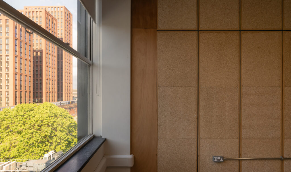 A window view of tall orange buildings and green trees in Manchester, with the interior wall featuring cork panels—a nod to Sustainable Ventures—and an electrical socket on the right side.