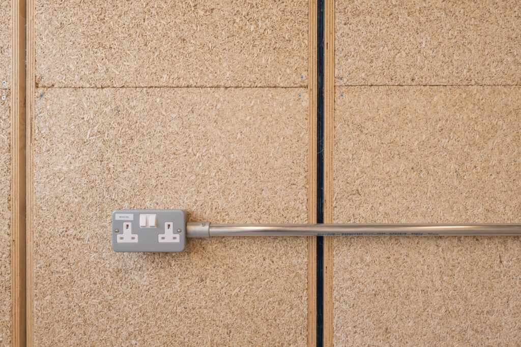 A double electrical socket with two switches is mounted on a light brown, textured wooden wall at Sustainable Ventures, Manchester, with a metal conduit running horizontally from the socket.