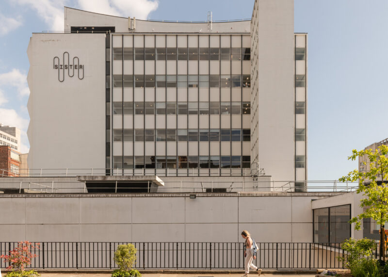 A person walks along a pavement in front of a modern, multi-storey building with large windows and a sign reading Sister on the upper left side, under a blue Manchester sky—home to innovative Sustainable Ventures.