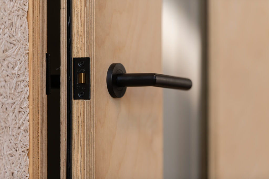 A close-up of a light wooden door slightly ajar, showing a black door handle and part of the metal latch plate on the door frame, typical of the modern Meeting Pods found in the Renold Building.