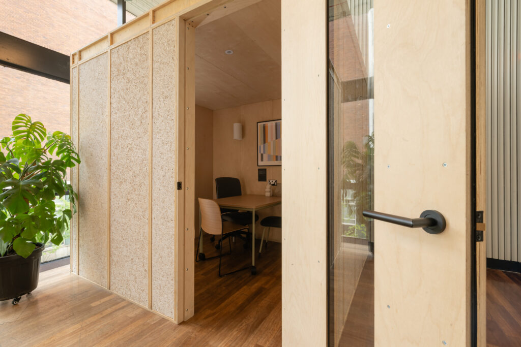 Small modern meeting room with wooden walls, a glass door, two chairs at a table, a plant outside, and a painting on the wall—all flooded with natural light—resembling the stylish Meeting Pods found in the Renold Building.