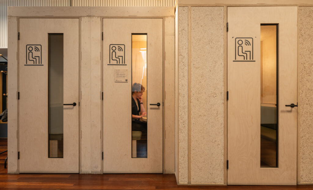 A person sits inside a small, private Meeting Pod with a glass-panelled door, talking on the phone. The pod is one of several lined up in the modern Renold Building, each marked with a conversation icon.