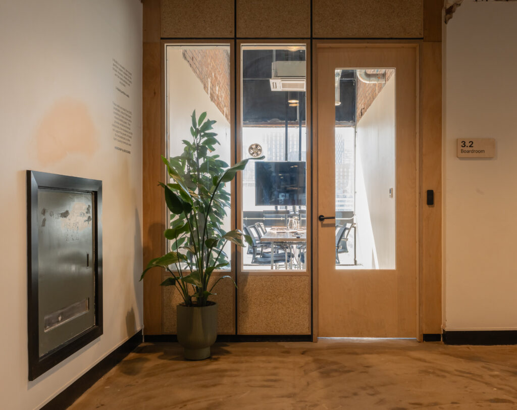 A modern office corridor at Sustainable Ventures Manchester features a potted plant near a wooden door labelled 3.2 Boardroom. Through the glass panels, a conference table, chairs, and TV screen can be seen inside the meeting room.