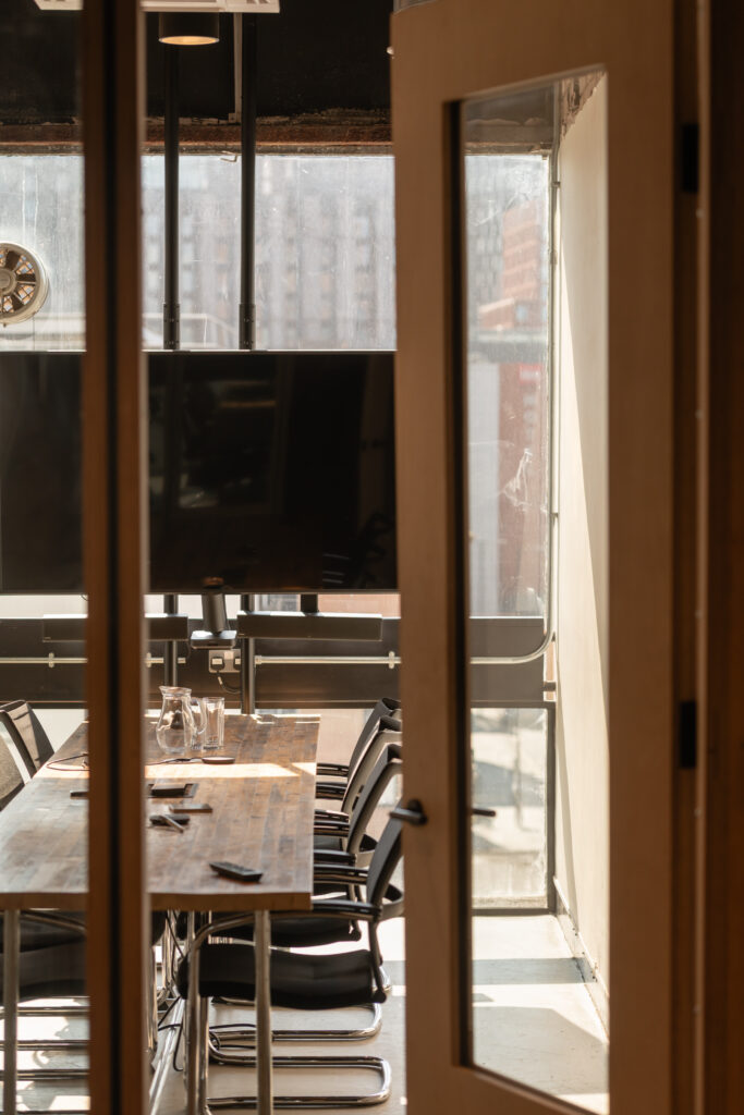 A sunlit meeting room in Manchester, designed for sustainable ventures, features a long wooden table, several black chairs, glass jugs and glasses, and a television on the wall, all viewed through a partially open door.