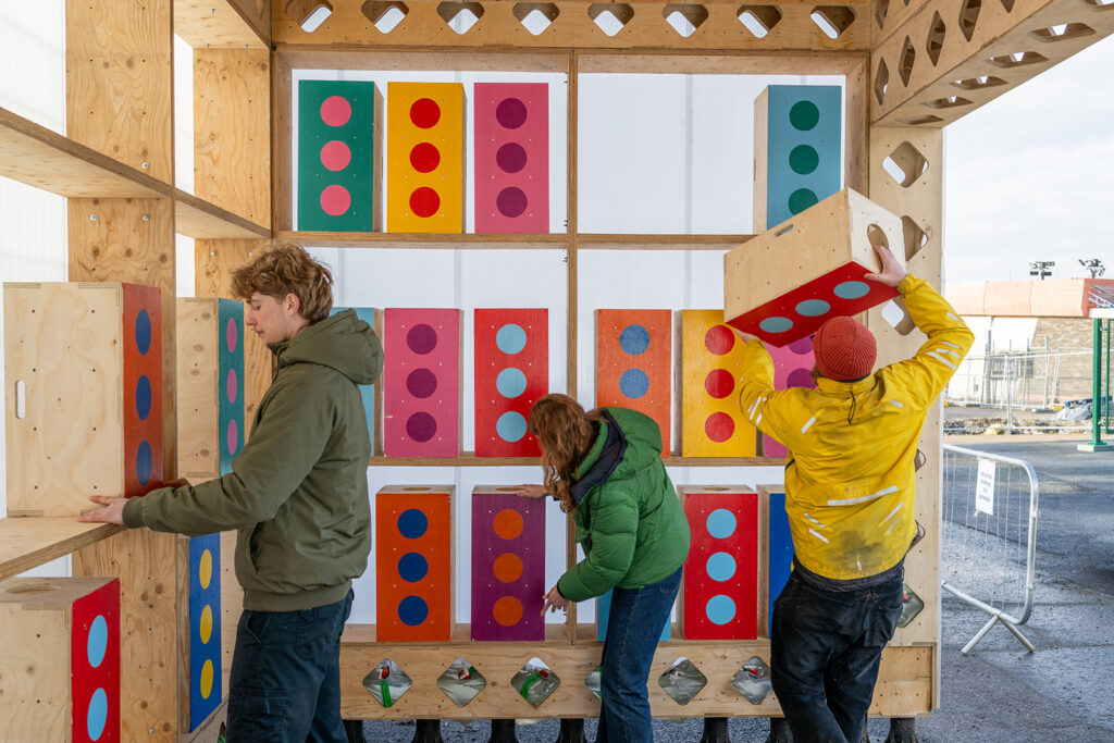 Three people arrange and stack featured, large, colourful blocks with polka dots on a wooden structure outdoors. Each block is painted with different coloured backgrounds and dots in what appears to be a lively public or community space.