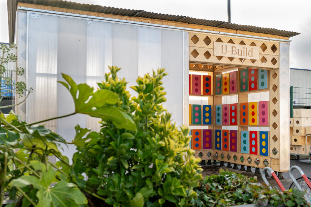 A modern wooden structure labelled U-Build, featured with colourful patterned panels, stands behind green leafy plants in an outdoor setting. The design showcases geometric shapes and a partially translucent wall.