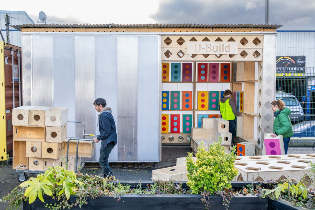 Three people move colourful modular panels and wooden boxes around a small wooden structure labelled “U-Build,” featured outdoors with plants in the foreground and industrial buildings in the background.
