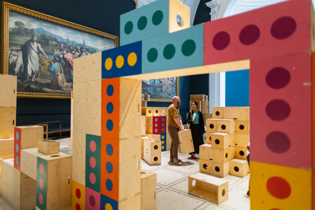 Two people stand among large, colourful wooden blocks in a gallery space at the V&A, surrounded by classical paintings on the walls. Engaging with the interactive Upstart exhibit, they collaborate to build structures for activism, creating a dynamic dialogue between art and action.