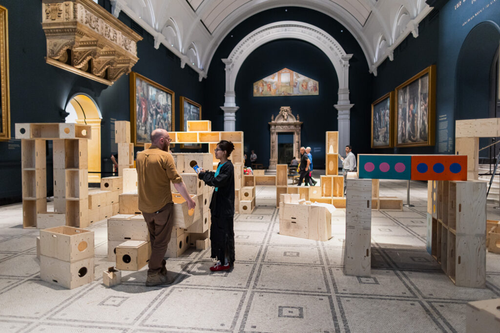 People interact in a museum gallery filled with large wooden building blocks, using them to build structures that spark creativity and collaboration. Decorative arches, artwork, and a tiled floor are visible, with adults and children scattered throughout the space.