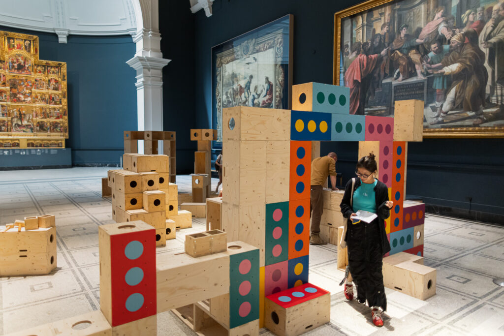 A person walks through a V&A museum gallery with large paintings on the walls and colourful wooden blocks arranged in structures across the floor, reflecting efforts to build structures for activism and upstart creativity.