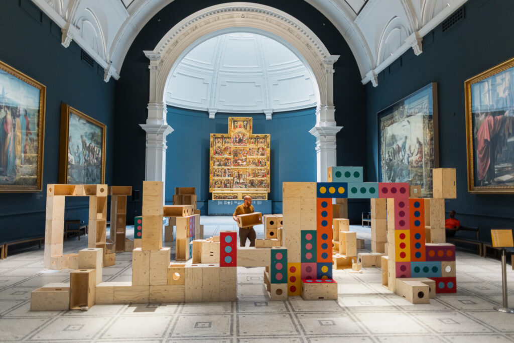 A person stands among large wooden blocks with colourful dots, arranged as a playful installation in the grand V&A museum gallery. Surrounded by classical paintings and an arched ceiling, the scene invites visitors to explore how creativity and upstart ideas can build structures for activism within this iconic space.