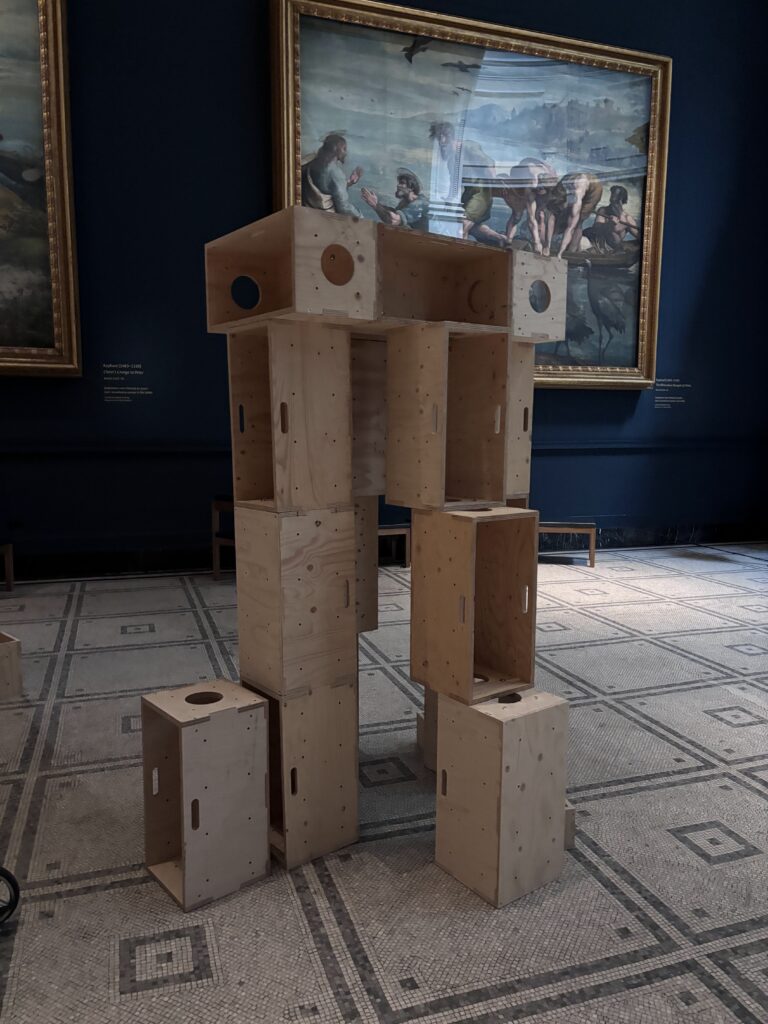 A sculpture made of stacked wooden crates stands in a gallery at the V&A, surrounded by classical paintings on the blue walls and a patterned tiled floor—a striking example of how upstart artists build structures for activism within traditional spaces.