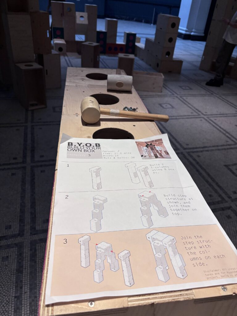 A wooden workbench with holes, a mallet, and printed step-by-step construction instructions—part of the Upstart initiative at the V&A—invites visitors to build structures for activism; wooden blocks and structures are visible in the background on a tiled floor.