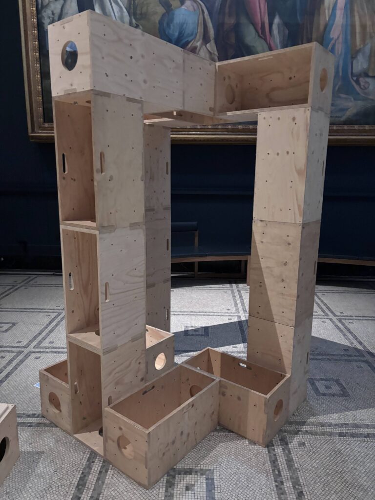 A structure made of stacked, unfinished wooden boxes with holes is displayed indoors on a tiled floor, reminiscent of the creative initiatives showcased at the V&A. A framed painting in the background complements this thought-provoking Upstart installation, inspiring viewers to build structures for activism through innovative design.