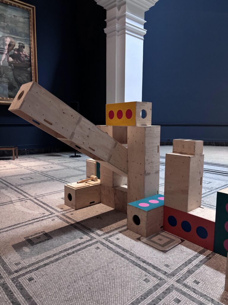Wooden blocks of various sizes and colours are stacked into a playful, abstract structure in the V&A art gallery, where tiled floors and classical architectural details set the stage for visitors to explore how we build structures for activism in contemporary spaces.