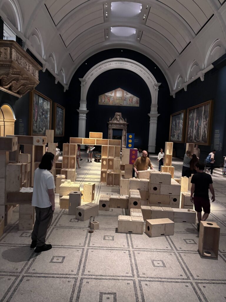 People interact with large wooden blocks arranged in a creative structure inside the grand halls of the V&A, where arched ceilings and classical paintings provide an inspiring backdrop. This immersive installation encourages visitors to build structures for activism, sparking dialogue and engagement within the museum’s historic setting.
