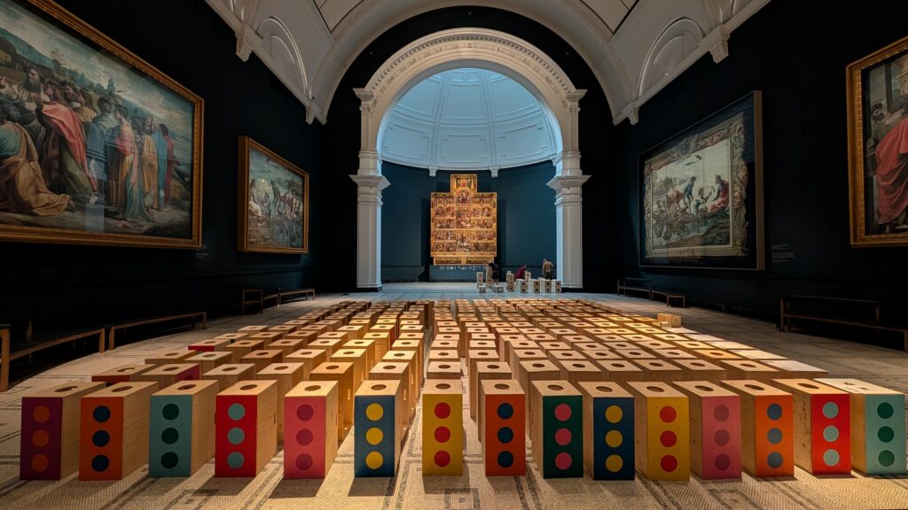 A grand museum hall in the style of the V&A, adorned with classical paintings and an ornate altar, features colourful rectangular blocks with circular patterns—an installation that invites visitors to build structures for activism—carefully arranged in rows on the floor.